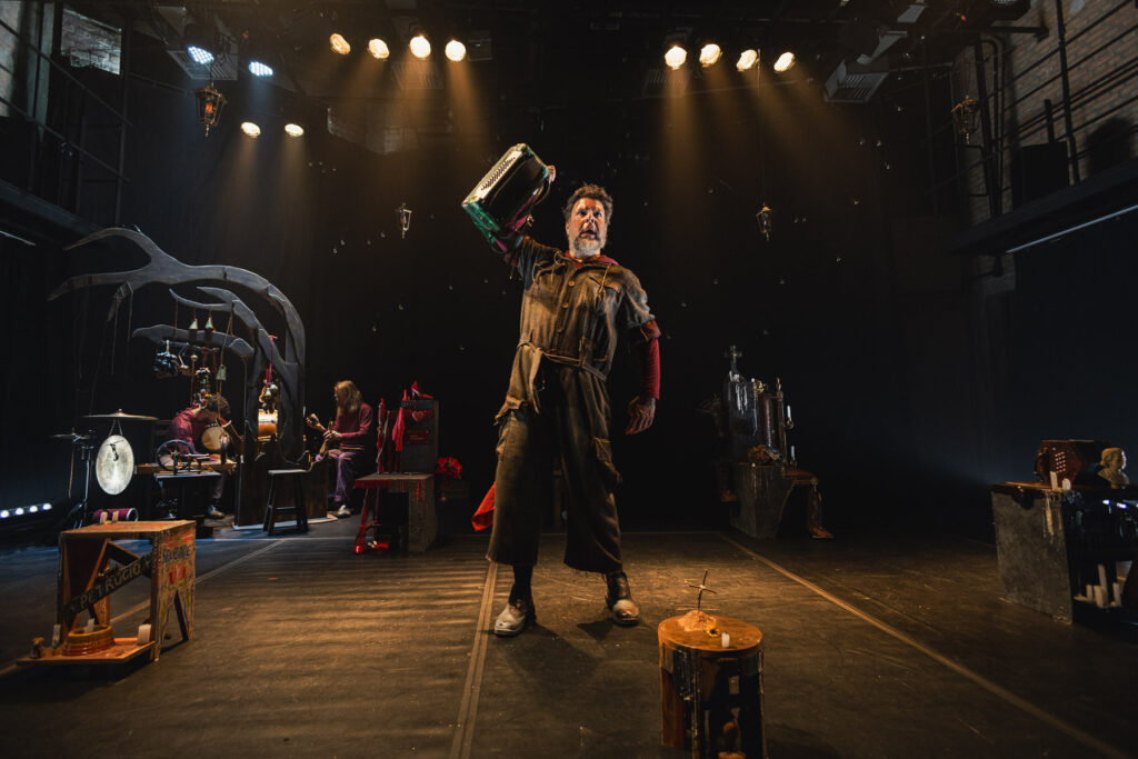 Um ator em vestuário cénico ergue uma concertina com uma mão no centro de um palco de teatro iluminado por projetores, cercado por instrumentos musicais e elementos decorativos.