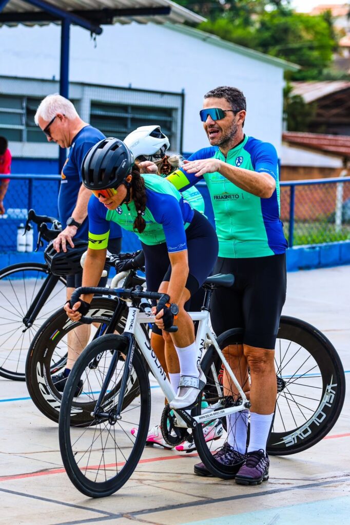 O treinador Marcos Novello surge ao lado de uma ciclista equipada com capacete e óculos de sol, orientando-a na posição de partida sobre a sua bicicleta num velódromo.