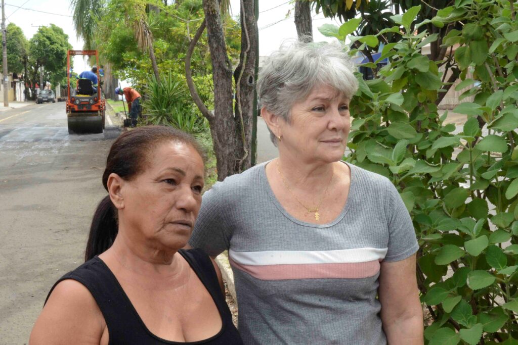 Duas moradoras em primeiro plano observam os trabalhos de manutenção na rua, onde se vê um rolo compressor e um trabalhador ao fundo.