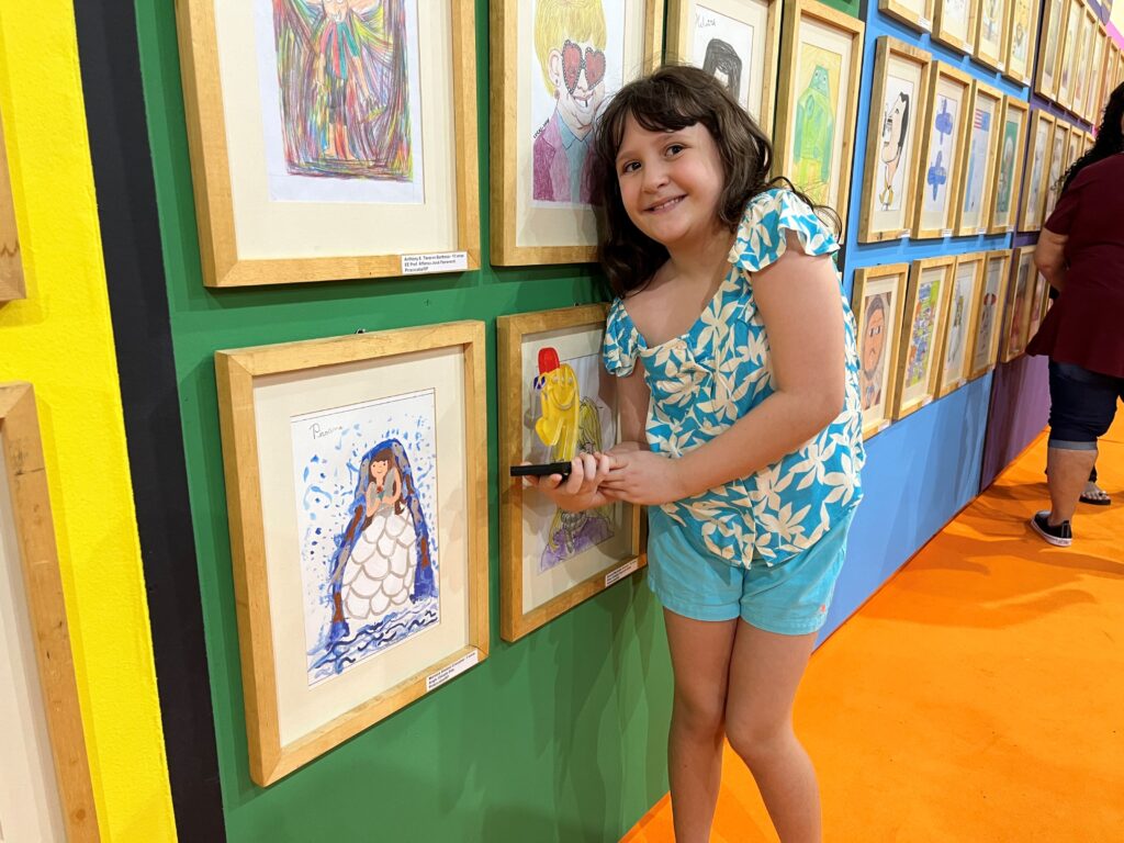 Mariana Simões Crocomo sorri ao lado do seu desenho premiado com o 1.º lugar na categoria de 7 a 10 anos.