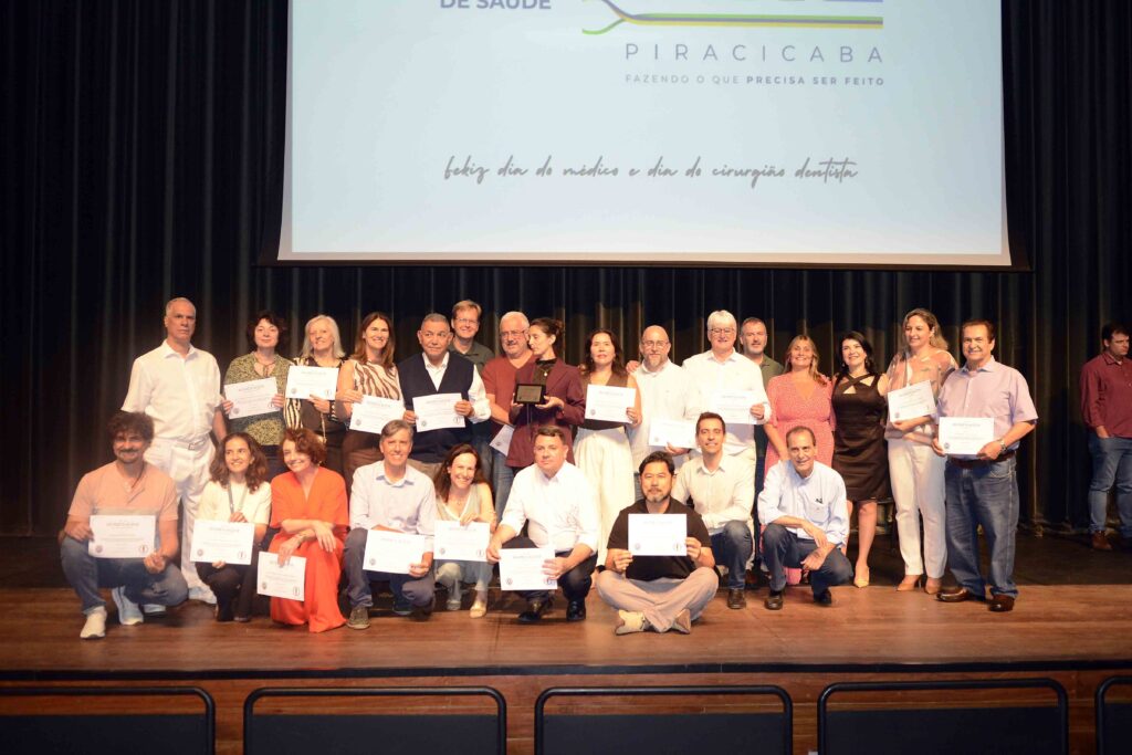Grande grupo de médicos e cirurgiões-dentistas homenageados reunidos no palco, exibindo os seus certificados de reconhecimento diante da tela do evento.