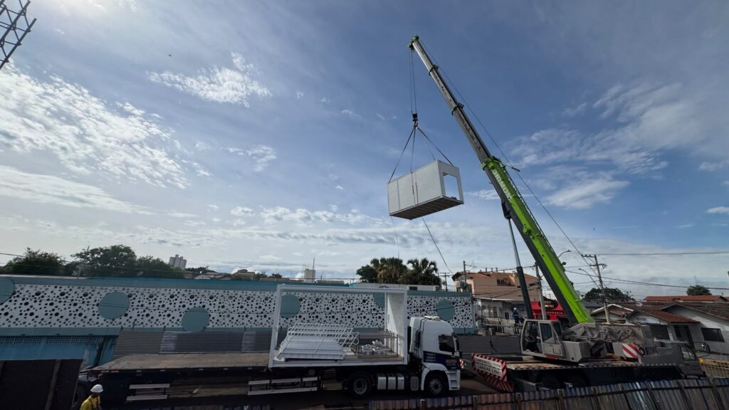 Uma grande autogrua telescópica verde e cinzenta ergue um módulo pré-fabricado no ar para o posicionar na obra, a partir de um camião de transporte estacionado junto ao edifício.