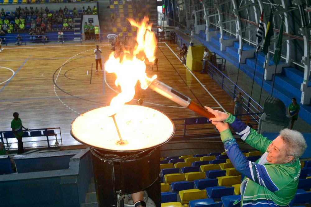 A atleta Leila Marta Rodrigues Gomes (Jacaré) segura a tocha e acende a pira olímpica com chamas acesas, posicionada nas bancadas do ginásio.