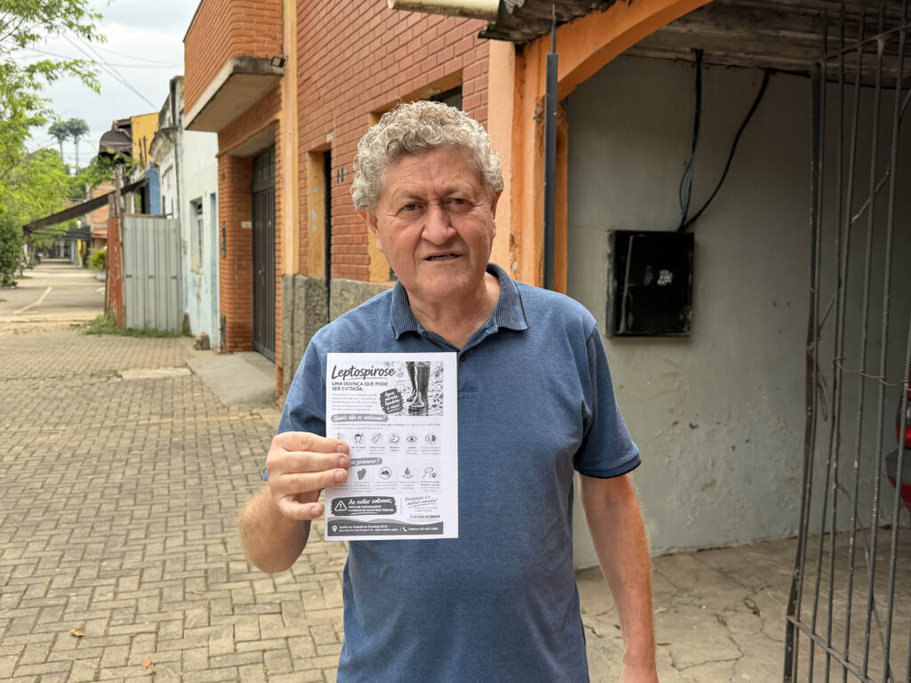 Um homem de cabelo grisalho e t-shirt azul em pé na rua, a segurar um folheto informativo com orientações e prevenção sobre a leptospirose.