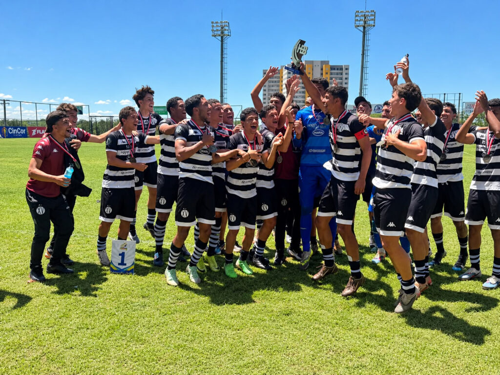 Um time de futebol juvenil celebra entusiasticamente a conquista do primeiro lugar em um campo gramado e ensolarado. Eles vestem uniformes listrados em preto e branco (estilo XV de Piracicaba), exibem medalhas e levantam o troféu. Há uma placa com o número '1' em destaque