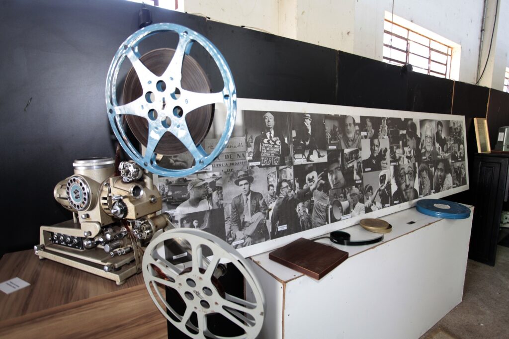 Uma exibição museológica com um projetor cinematográfico antigo de bobinas abertas, acompanhado por um painel expositivo com fotografias a preto e branco e outros acessórios audiovisuais dispostos numa bancada.