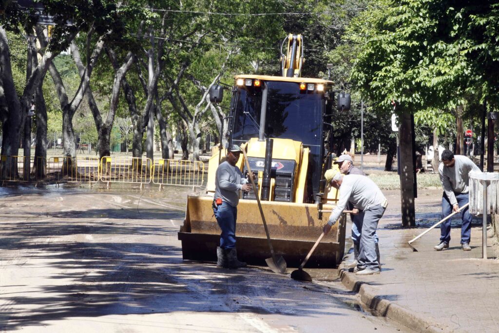 Quatro trabalhadores com ferramentas manuais (pás e rodos) limpam a lama acumulada no chão e no meio-fio de uma avenida arborizada, logo à frente de uma retroescavadeira amarela posicionada na pista.