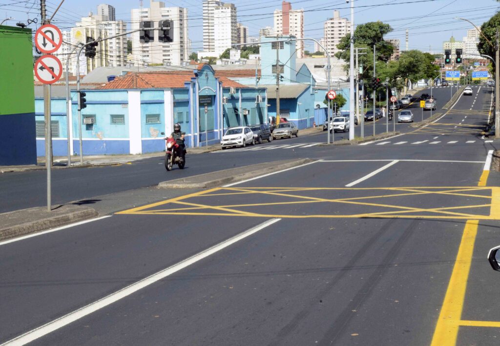 Vista de uma avenida ampla com o asfalto novo totalmente recapeado, acompanhado por sinalização horizontal recente (linhas brancas e amareladas). Ao fundo, observam-se edifícios da cidade, prédios de cor azul e um motociclista a circular na via.