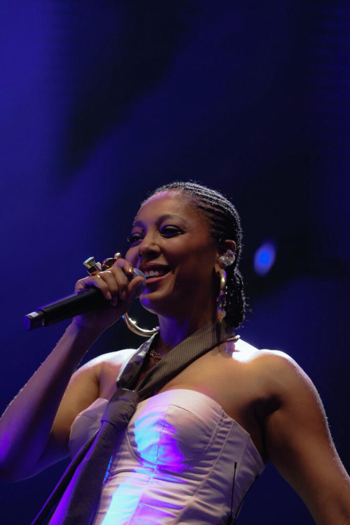 A cantora Negra Li sorri enquanto canta num microfone durante uma apresentação ao vivo, vestindo um corset claro com gravata e iluminação azul de fundo.