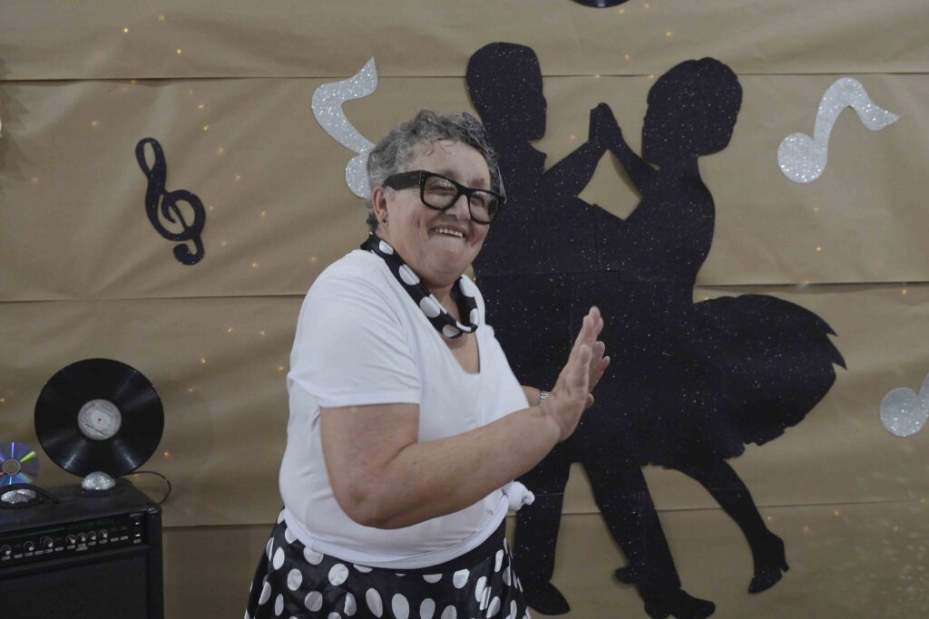 Uma idosa de óculos e saia de bolinhas dança sorridente em frente a um painel temático decorado com notas musicais e silhuetas de um casal a dançar.