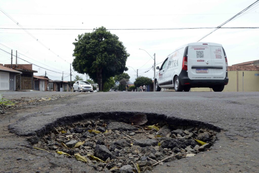 Buraco profundo e desgastado no asfalto em primeiro plano, com uma carrinha de entregas branca a passar ao fundo.