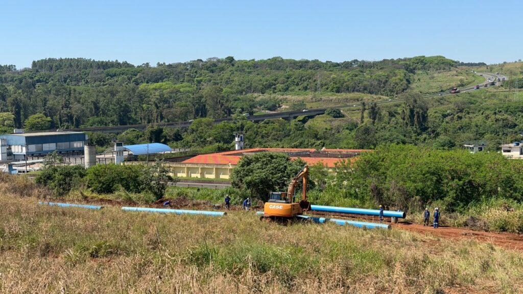 Vista ampla das obras de uma adutora em meio à vegetação. Trabalhadores e uma escavadeira atuam na montagem da rede de tubulações, com rodovia e construções ao fundo.
