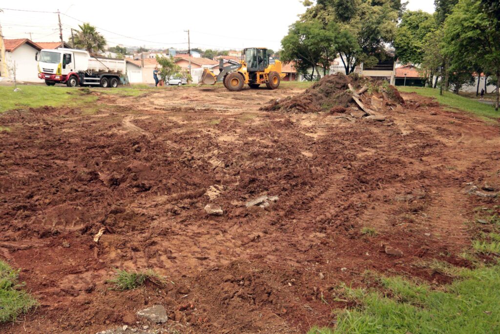 Um terreno em obras com terra batida e revolvida, onde uma pá carregadora amarela opera ao fundo ao lado de um camião basculante branco e pilhas de terra acumulada.
