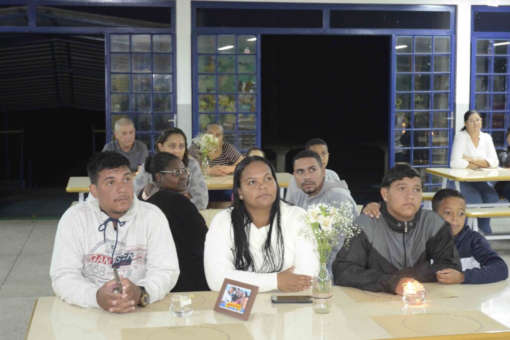 Participantes das famílias reunidas assistem à apresentação sentados em mesas decoradas no pátio da escola