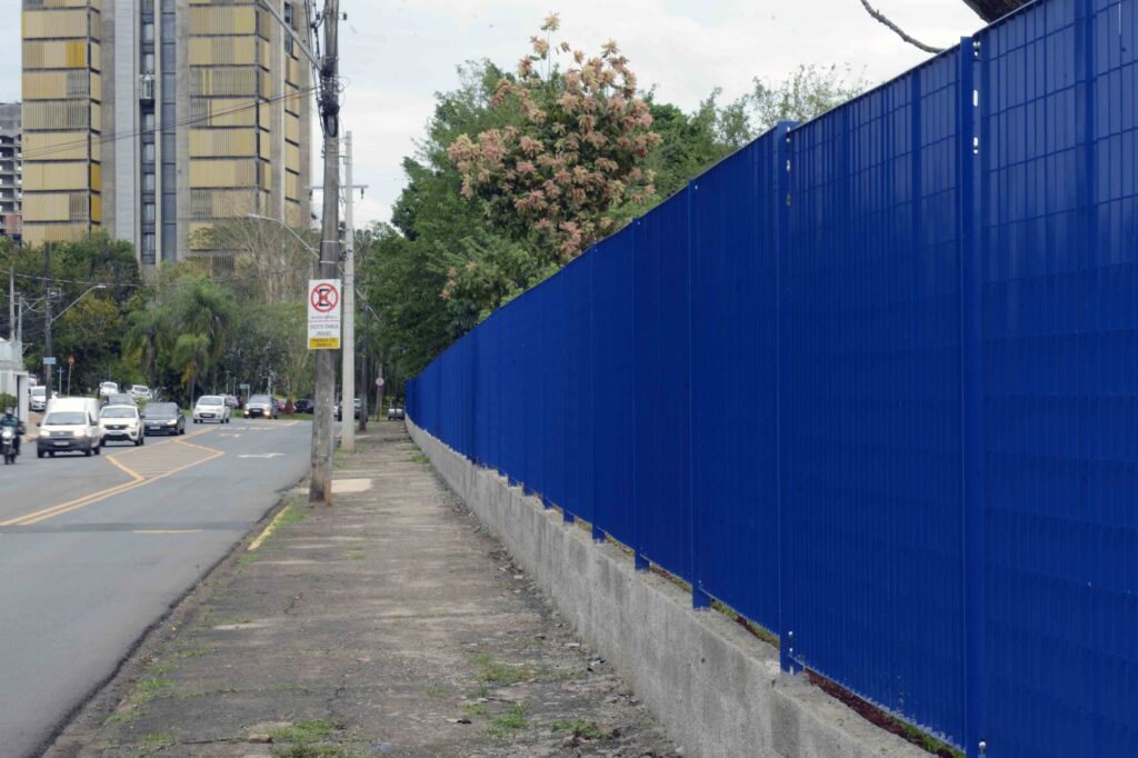 Vista lateral de um novo gradil azul de proteção montado sobre uma mureta de betão, ao longo do passeio de uma avenida movimentada.