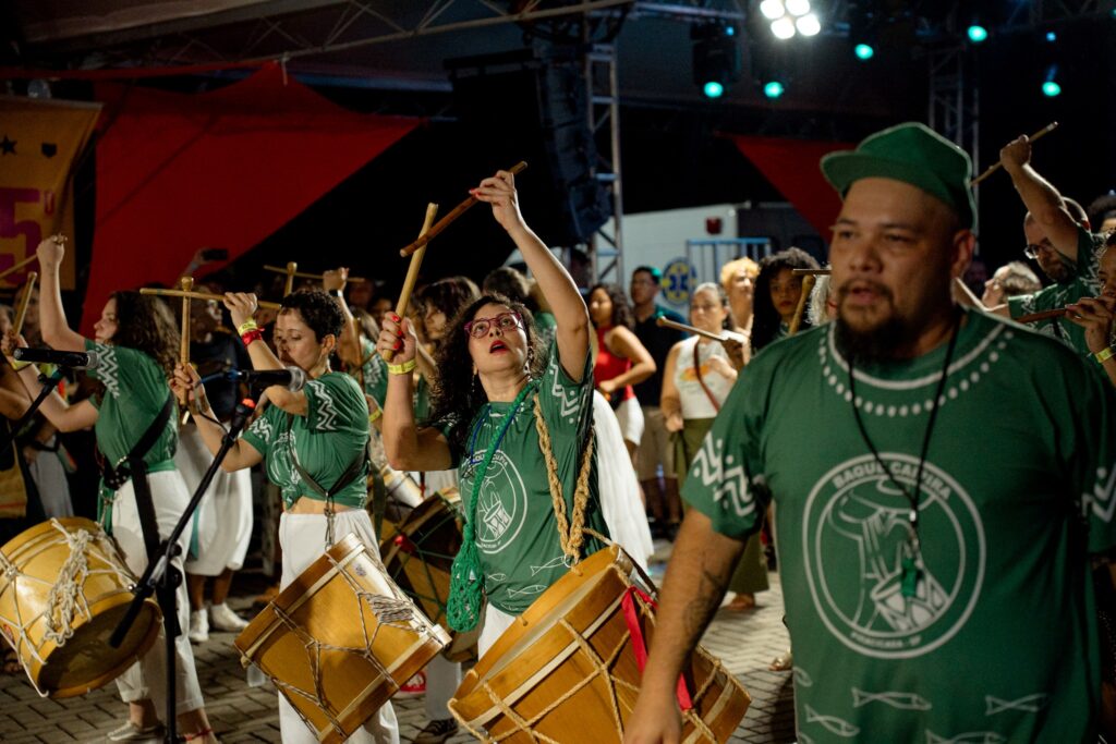 Integrantes do grupo Baque Caipira vestidos com camisas verdes e calças brancas, a tocar tambores e alfaia durante o Festival Curau.