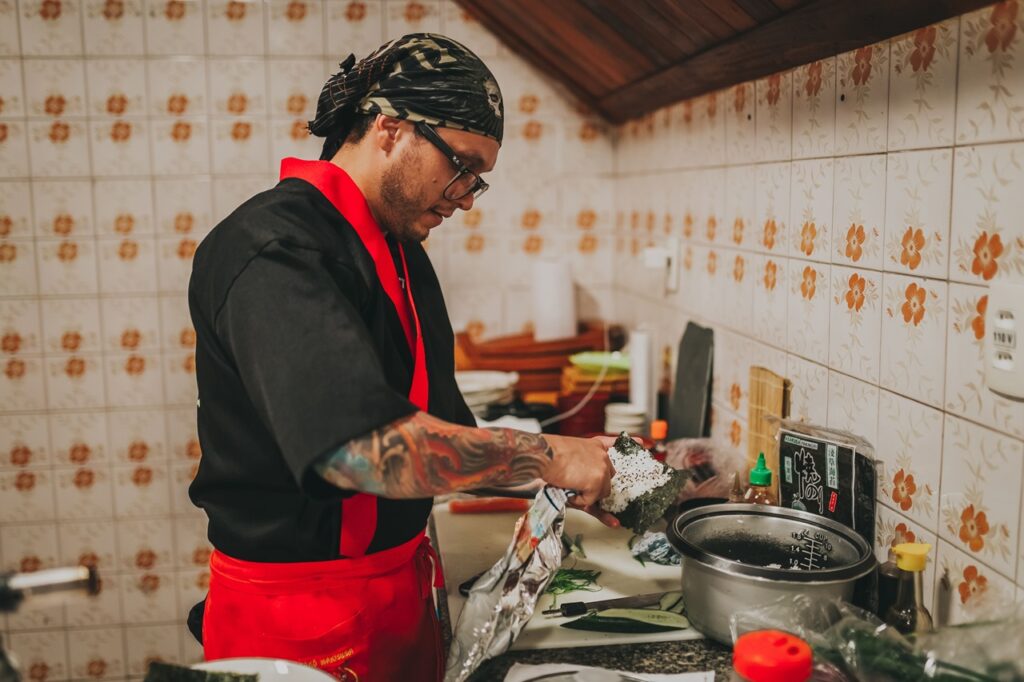 Cozinheiro com dólmã preto e lenço na cabeça a preparar temaki de culinária japonesa numa bancada de cozinha.