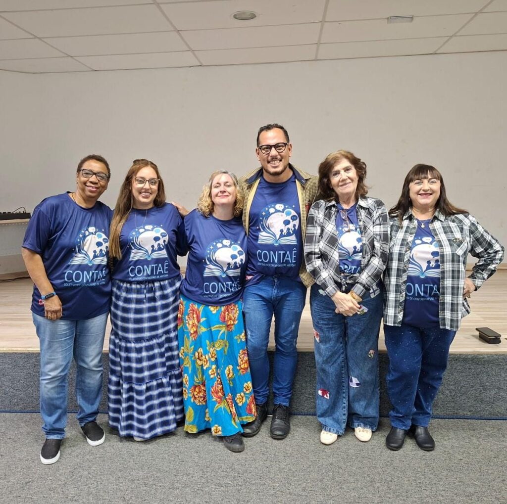 Membros do coletivo "Contaê" posam lado a lado sorridentes num palco, todos vestindo a t-shirt azul do grupo de contação de histórias.