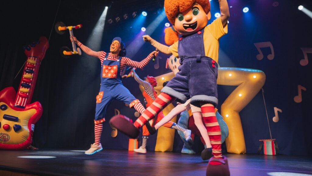 A personagem Mika dá um salto animado em primeiro plano no palco, acompanhada por atores vestidas de palhaços e outras personagens fantasiadas numa cena vibrante do espetáculo infantil.