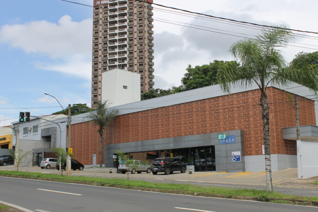 Fachada da sede do Ipasp, edifício moderno com revestimento em tijolos vazados e estrutura cinzenta, com estacionamento frontal e um prédio residencial ao fundo.