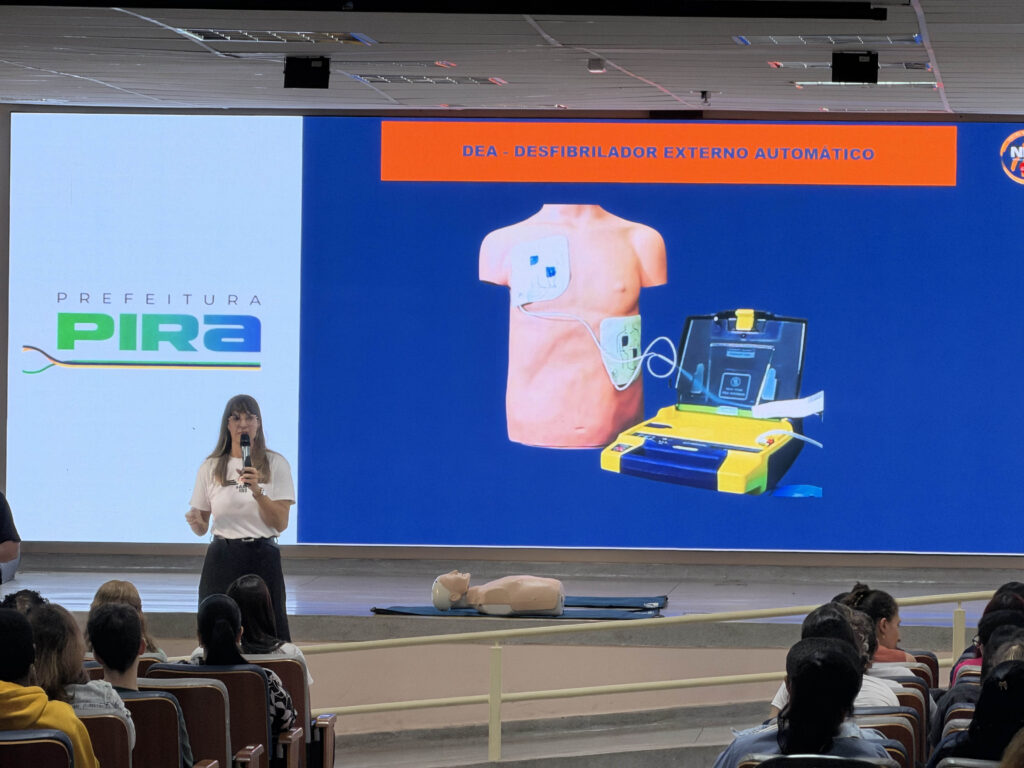 Uma palestrante no palco de um auditório a apresentar um slide explicativo sobre o uso do Desfibrilador Externo Automático (DEA), com a plateia de educadores a assistir de costas.