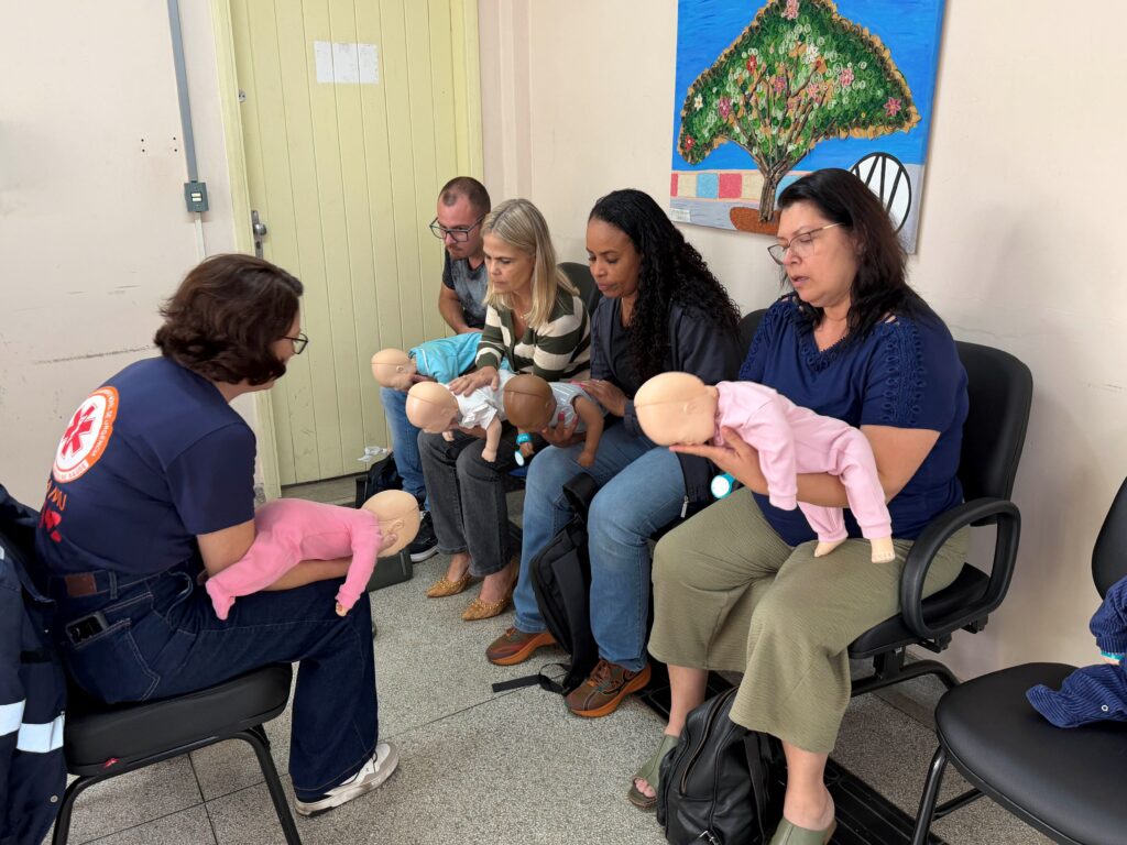 Uma instrutora do Samu sentada em frente a um grupo de educadores, demonstrando na prática as técnicas de desobstrução de vias aéreas em manequins de bebês.