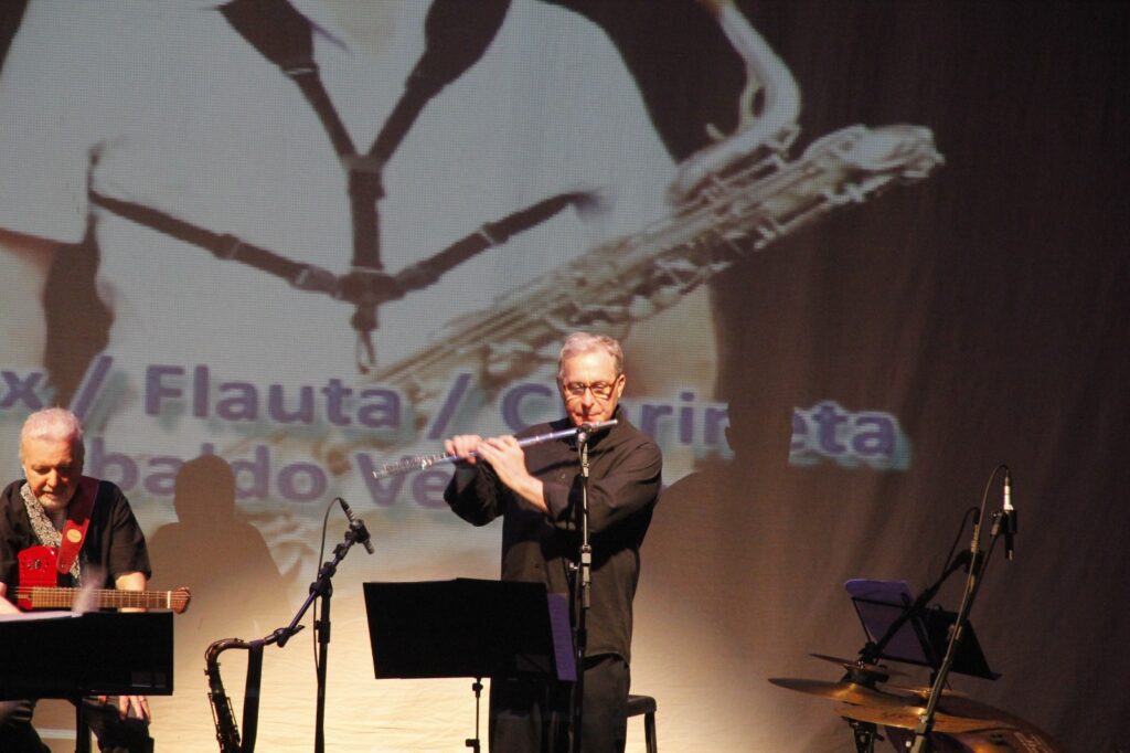 O músico Ubaldo Versolato toca flauta transversal no palco do Teatro Erotides de Campos durante a apresentação do projeto "Por Trás da Canção", acompanhado por outro instrumentista ao fundo.