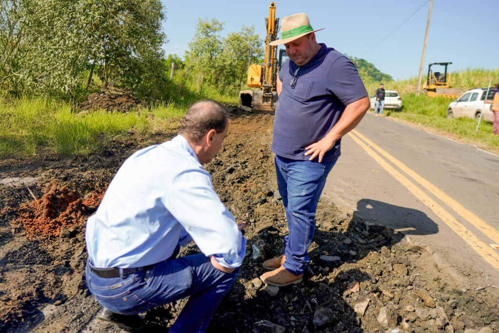 O prefeito Helinho Zanatta inspeciona obras de manutenção numa estrada, acompanhado por um trabalhador, com máquinas pesadas ao fundo.