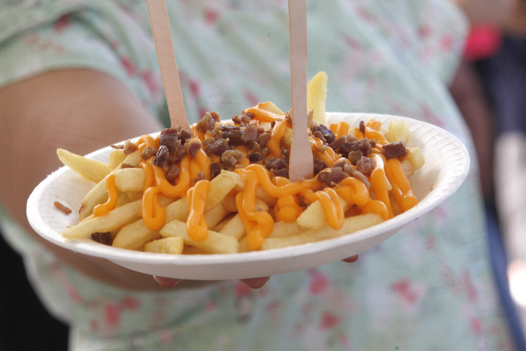Um prato descartável com batatas fritas cobertas de molho de queijo cheddar e pedaços de bacon, servido com dois garfos de madeira.