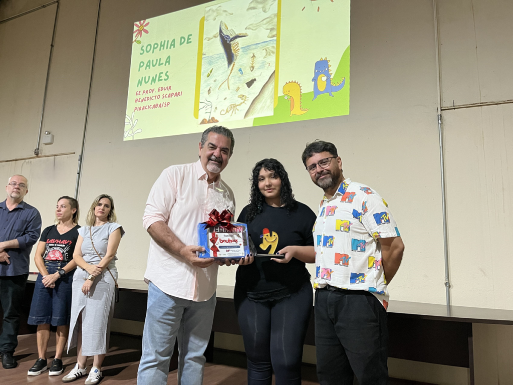 O Secretário de Cultura Carlos Beltrame e Junior Kadeshi (Cedhu) entregam o prémio e o troféu a Sophia de Paula Nunes no palco.