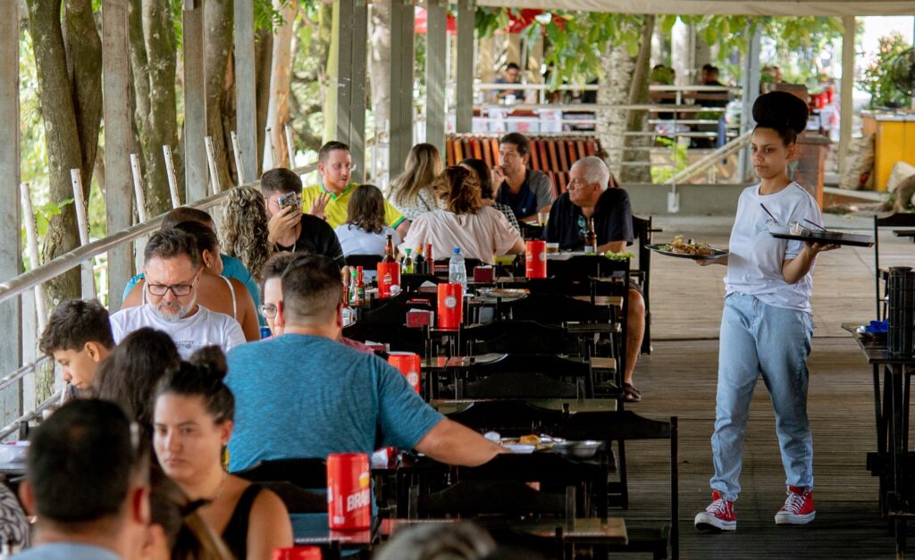 Uma empregada de mesa carrega uma bandeja enquanto caminha pelo corredor de um restaurante movimentado e coberto. Em primeiro plano e ao longo do salão, vários clientes estão sentados a mesas de madeira a almoçar e a conviver.