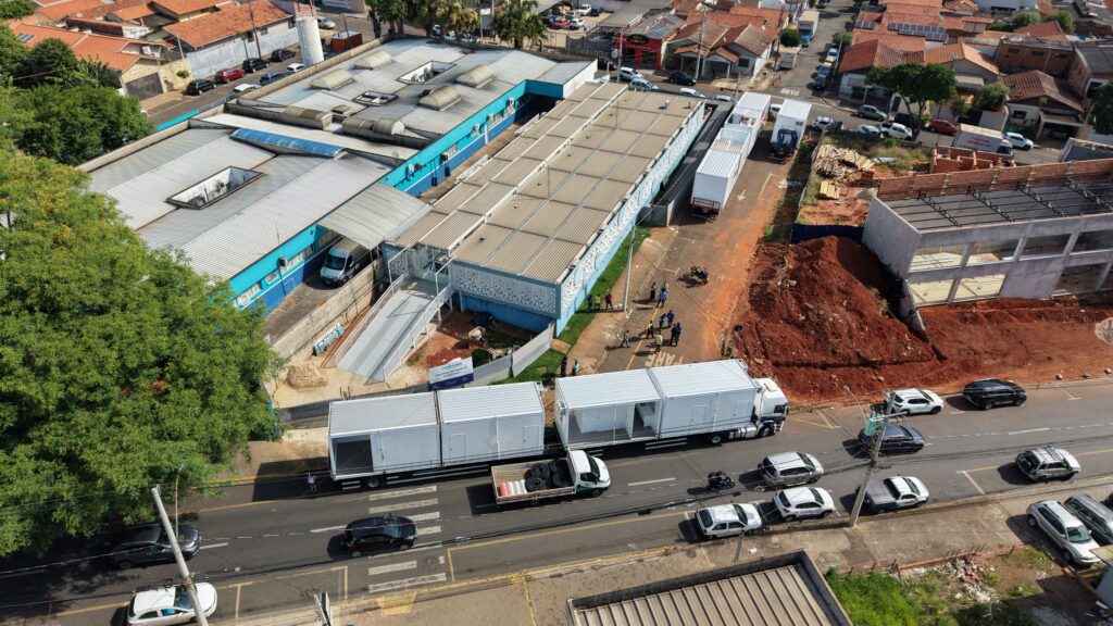 Uma vista aérea (feita por drone) mostra o complexo hospitalar e a rua envolvente, onde camiões de grande porte transportam os módulos pré-fabricados brancos para a obra de ampliação do prédio.