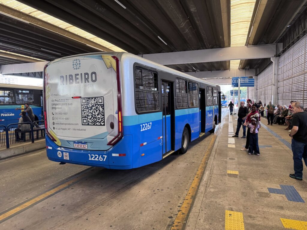 Um autocarro urbano azul e branco parado numa plataforma de um terminal rodoviário/urbano, com passageiros a aguardar ao longo da plataforma. A traseira do autocarro exibe um anúncio publicitário e o número de frota 12267.
