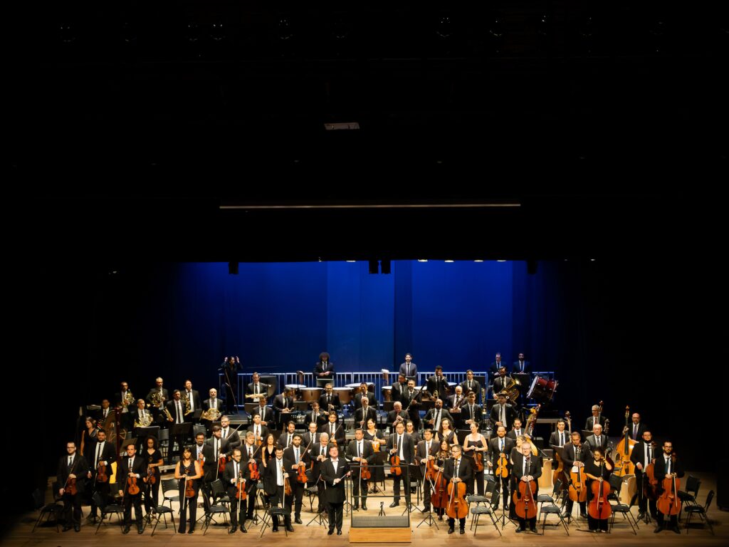 Apresentação da Orquestra Sinfônica de Piracicaba (OSP) no palco, com músicos vestidos de preto com os seus instrumentos e o maestro ao centro sob iluminação azul.