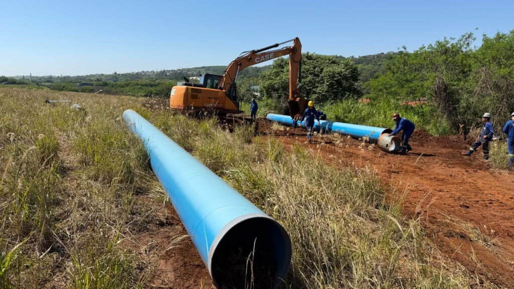 Obra de infraestrutura em área rural, com equipes trabalhando na instalação de grandes tubulações azuis. Uma escavadeira auxilia o posicionamento dos tubos ao longo de uma faixa de terra.