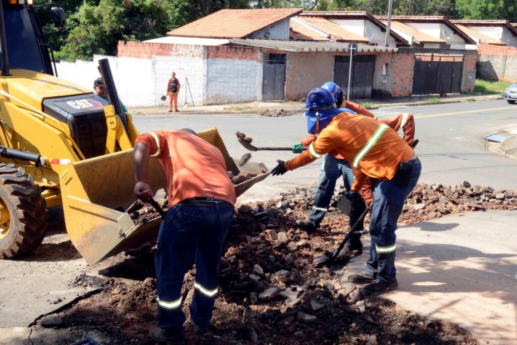 Trabalhadores equipados com capacetes, luvas e pás removem detritos de asfalto e preparam a pista, auxiliados por uma retroescavadeira amarela.