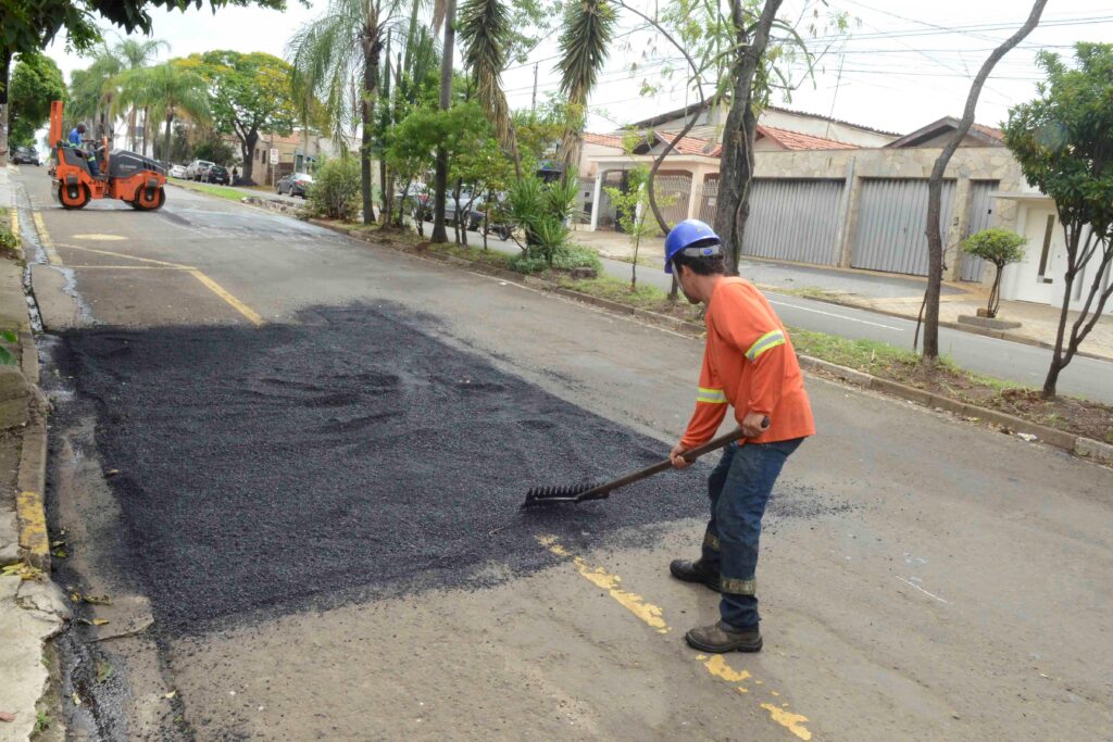 Um operário com equipamento de proteção espalha e nivela asfalto fresco durante uma operação de reparação de pavimentos numa via pública.