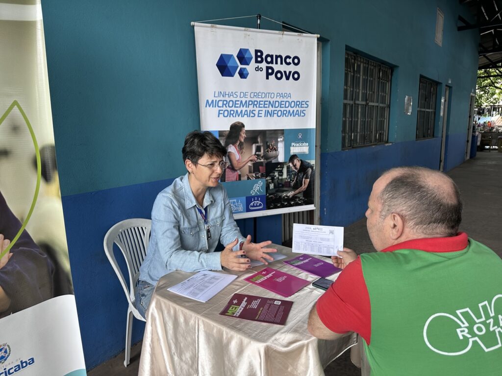 Uma orientadora atende um feirante numa mesa informativa do "Banco do Povo", prestando esclarecimentos sobre linhas de crédito e serviços para microempreendedores.