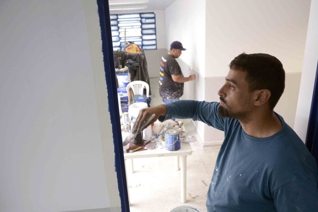 Mostra um pintor em primeiro plano com uma camisola azul a aplicar tinta no batente de uma porta, enquanto outro homem ao fundo pinta uma parede branca num espaço em obras cheio de latas de tinta e ferramentas.