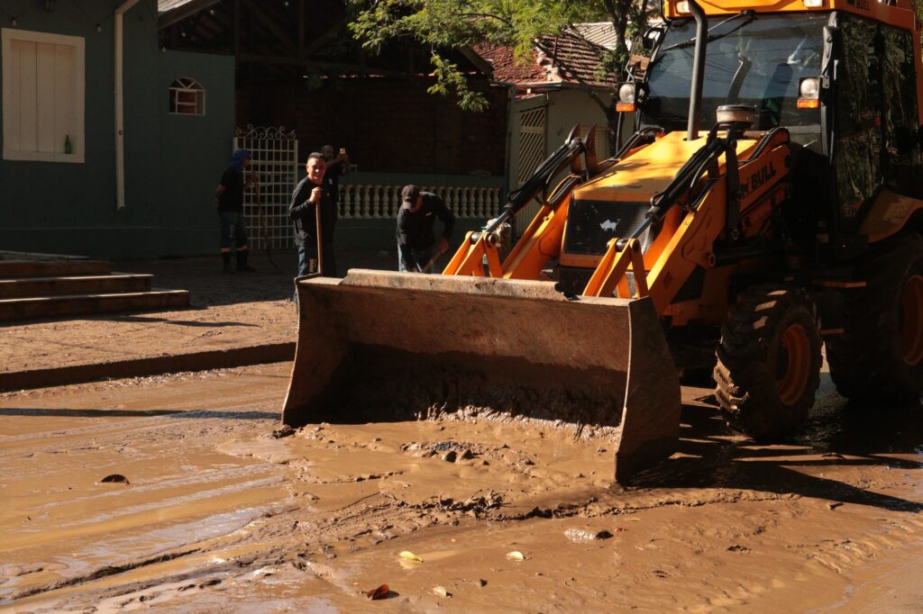 Mostra em primeiro plano a pá frontal de uma retroescavadeira amarela (Bull) a recolher uma espessa camada de lama acumulada na pista. Ao fundo, vários trabalhadores com pás auxiliam manualmente no processo de limpeza em frente a edifícios residenciais/comerciais.