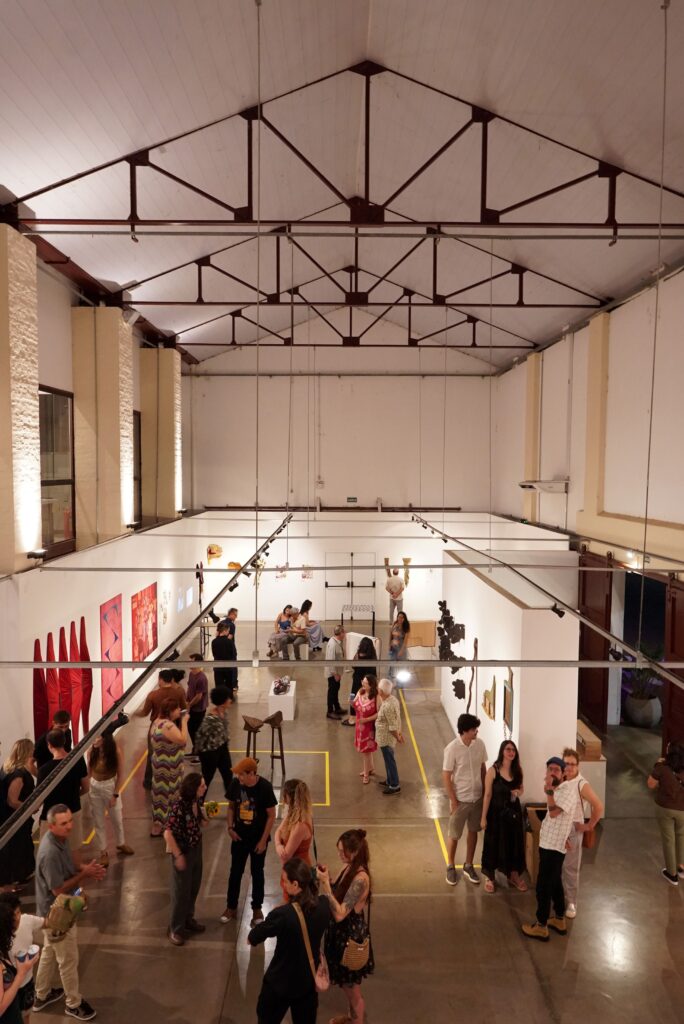 Mostra uma vista superior e panorâmica do interior de uma galeria de arte ou espaço de exposição com teto alto e vigas metálicas expostas. Diversas pessoas circulam, observam as obras expostas nas paredes e pedestais, ou conversam no amplo salão iluminado.