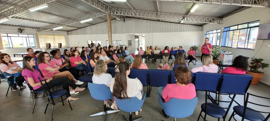 Perspetiva lateral e aproximada do mesmo encontro, mostrando as participantes sentadas em cadeiras dispostas em semicírculo. A palestrante em pé interage e conduz a conversa num ambiente amplo com grande cobertura de teto metálico.