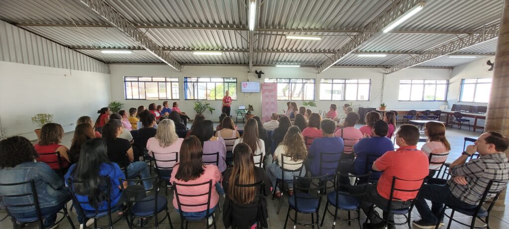 Uma vista do fundo de um auditório iluminado onde dezenas de servidoras e colaboradores acompanham uma apresentação sobre a prevenção do cancro de mama. À frente, a palestrante veste uma t-shirt cor-de-rosa perto de um ecrã e de uma faixa informativa.