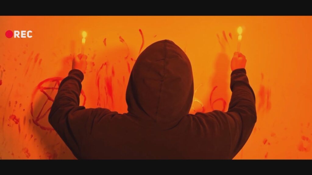 Fotograma de estilo found footage com iluminação alaranjada e um indicador "REC" no canto superior esquerdo. Mostra uma pessoa de costas com capuz escuro, a segurar velas acesas diante de uma parede com inscrições a vermelho, incluindo símbolos esotéricos e números.