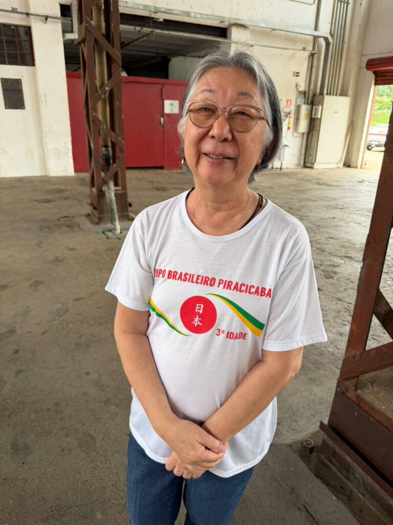 Uma mulher de óculos e cabelos grisalhos a sorrir para a câmara. Ela veste uma t-shirt branca do grupo "Nippo Brasileiro Piracicaba - 3ª Idade" e encontra-se num espaço amplo do Engenho Central.