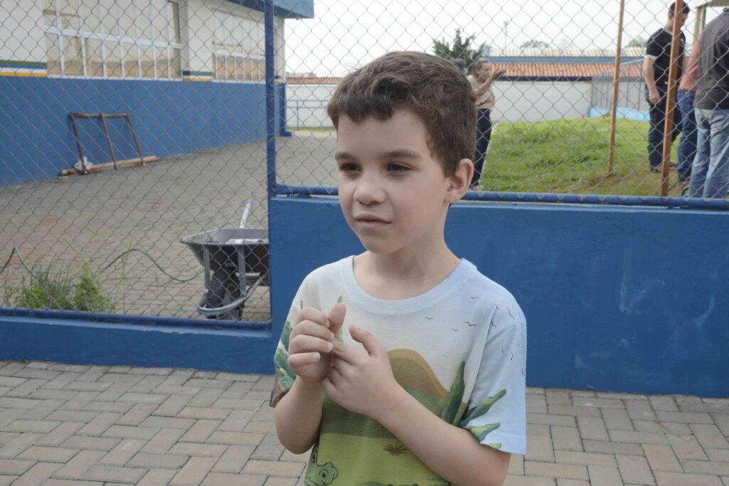 O estudante Fernando Ribeiro, de sete anos, veste uma t-shirt ilustrada e olha para o lado com uma expressão atenta e curiosa durante a atividade ao ar livre na escola.