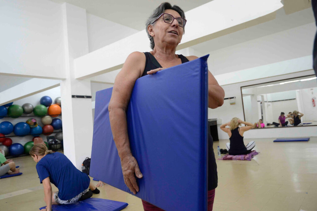 Mulher idosa em pé numa sala de ginástica segura um colchonete azul dobrado, enquanto outros alunos praticam exercícios no chão ao fundo.