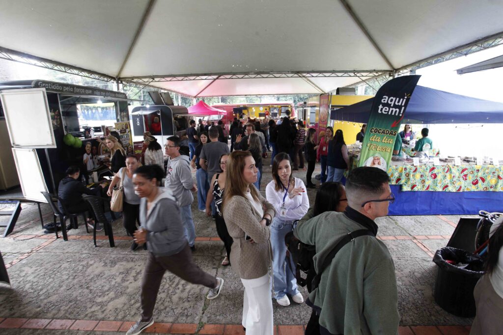 Um grupo de pessoas circula sob uma tenda exterior onde estão montadas várias unidades de food trucks e bancas comerciais.