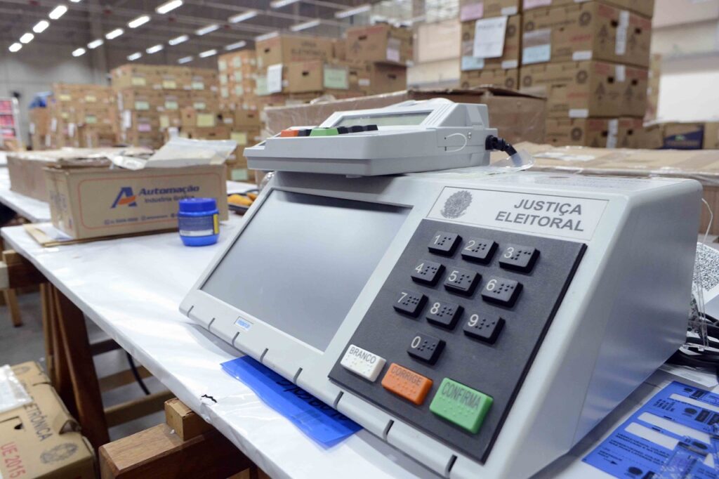 Um plano em detalhe de uma urna eletrónica da Justiça Eleitoral sobre uma mesa, destacando o teclado numérico, os botões de votação (Branco, Corrige e Confirma) e o ecrã. Ao fundo, observam-se várias caixas de papelão num depósito ou centro de logística.
