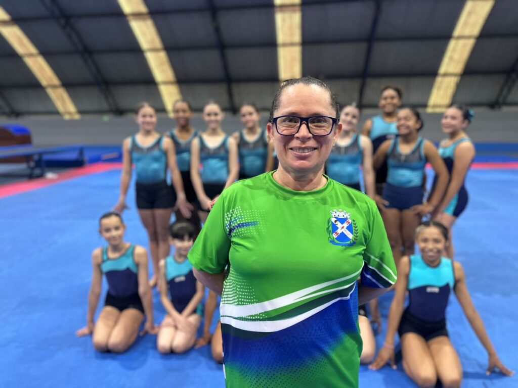 A professora Maria Dilailça sorri em primeiro plano com uma camisola verde oficial, acompanhada ao fundo por jovens atletas da equipa de ginástica acrobática vestidas com trajes azuis.
